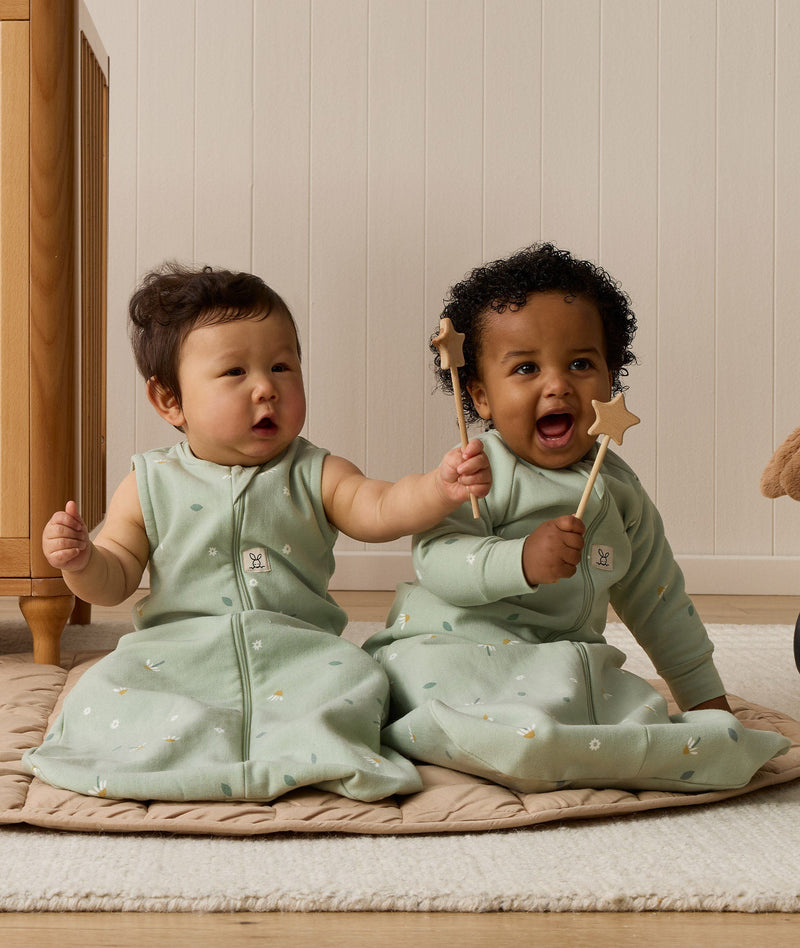 Two infants are sitting side-by-side while holding a wooden toy. One is wearing a sleeveless Sleeping Bag, while the other is wearing the same print with sleeves.