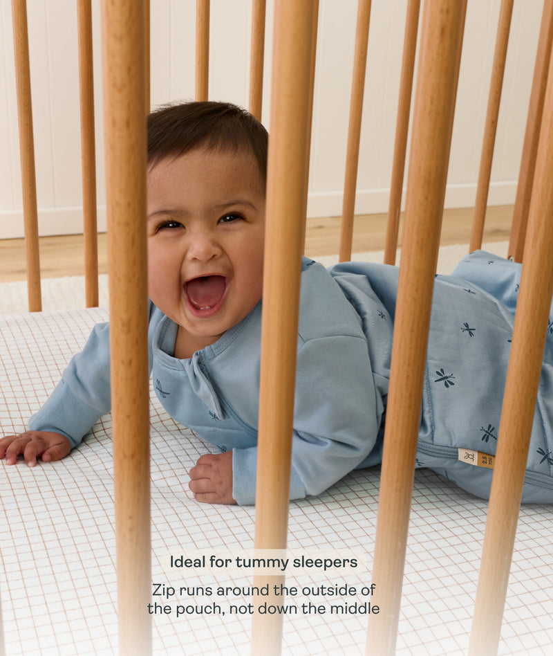 Baby lying on their tummy in a wooden cot wearing a blue long sleeved Sleeping Bag, with text noting it is ideal for tummy sleepers.
