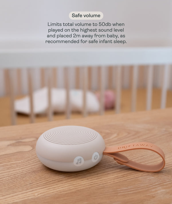 A compact, circular Drift Away white noise machine with a carry strap rests on a wooden surface. In the softly blurred background, a baby sleeps safely in a cot. Text overlay titled 