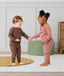 Two toddlers standing and holding hands indoors, wearing long-sleeve pyjama sets with matching pants—one in brown and one in dusty pink—designed for comfort and movement.