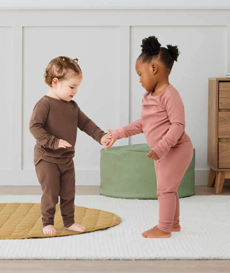 Two toddlers standing and holding hands indoors, wearing long-sleeve pyjama sets with matching pants—one in brown and one in dusty pink—designed for comfort and movement.
