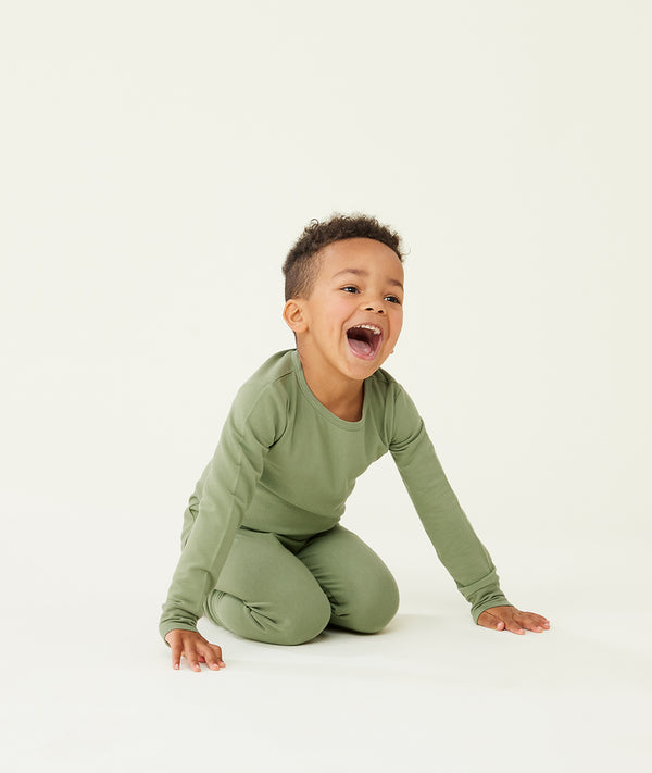 Preschooler kneeling on a cream background while wearing an olive green long-sleeve pyjama set with matching pants, designed for comfort and movement.