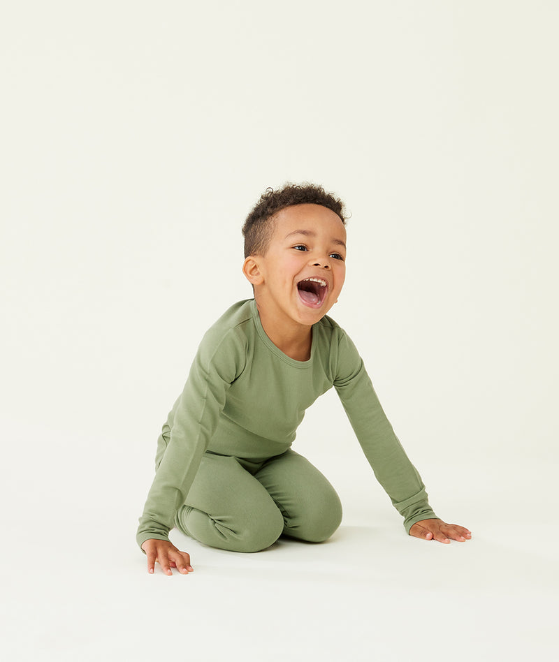 Preschooler kneeling on a cream background while wearing an olive green long-sleeve pyjama set with matching pants, designed for comfort and movement.