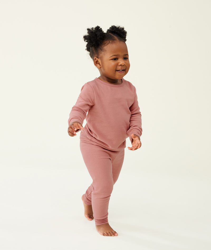 Toddler walking barefoot on a cream background wearing a blush pink long-sleeve pyjama set with matching pants, made from soft, comfortable fabric.