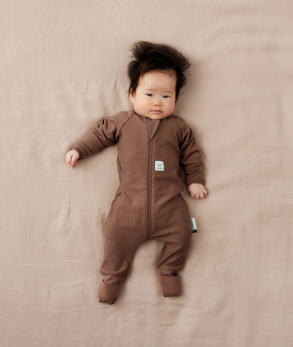 Baby lying on a beige blanket wearing a brown Long Sleeve Layer by ergoPouch, featuring a front-zip design and fold over foot cuffs.