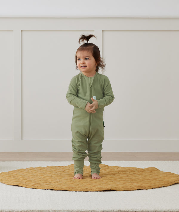 Toddler standing on a rug wearing an olive green Long Sleeve Layer by ergoPouch, made from soft, organic fabric.