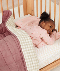 Toddler laying in bed on stomach, smiling, under ergoPouch Quilted Coverlet in Rosy Gingham