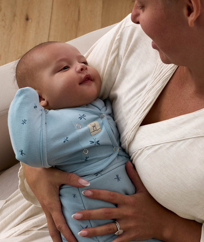 A mother holding a smiling baby in a light blue dragonfly-patterned Cocoon Swaddle Bag with Butterfly Cardi over top, looking down warmly at the infant.
