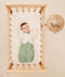An overhead view of a baby lying in a light wood bassinet, wearing a sage green Cocoon Swaddle Bag with a cream Butterfly Cardi layer over top, next to a small wooden side table with a white noise machine.