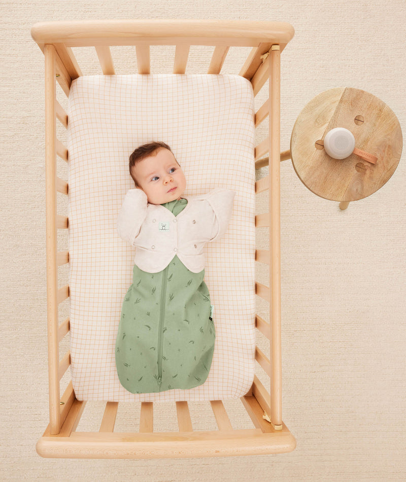 An overhead view of a baby lying in a light wood bassinet, wearing a sage green Cocoon Swaddle Bag with a cream Butterfly Cardi layer over top, next to a small wooden side table with a white noise machine.