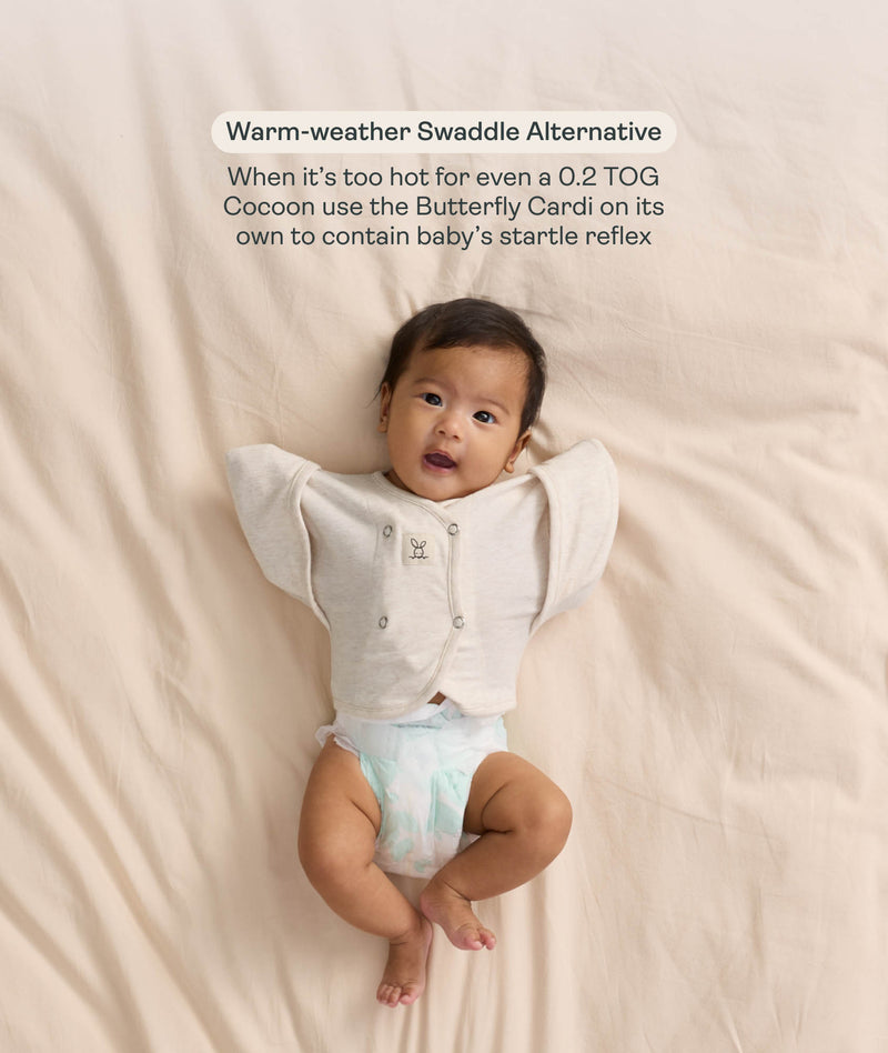 Baby in a nappy wearing a cream Butterfly Cardi, with text highlighting its use as a warm-weather swaddle alternative to help manage the startle reflex.