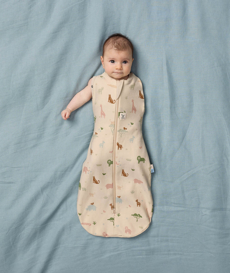A baby lies on a blue surface wearing a Cocoon Swaddle Bag with colourful Safari animal prints.