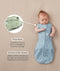 Baby lying on a beige surface in a blue dragonfly-pattern swaddle bag with arms out, featuring callout text highlighting press studs and a bell-shaped design.