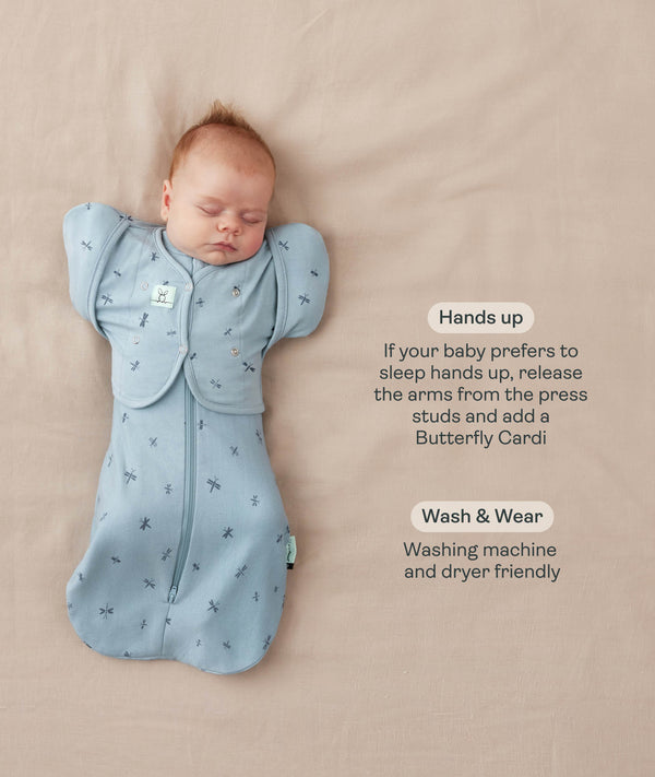 Baby lying on a beige surface in a blue dragonfly-pattern Cocoon Swaddle Bag paired with a Butterfly Cardi sleeve attachment, featuring product text callouts highlighting hands up sleeping and wash & wear care.