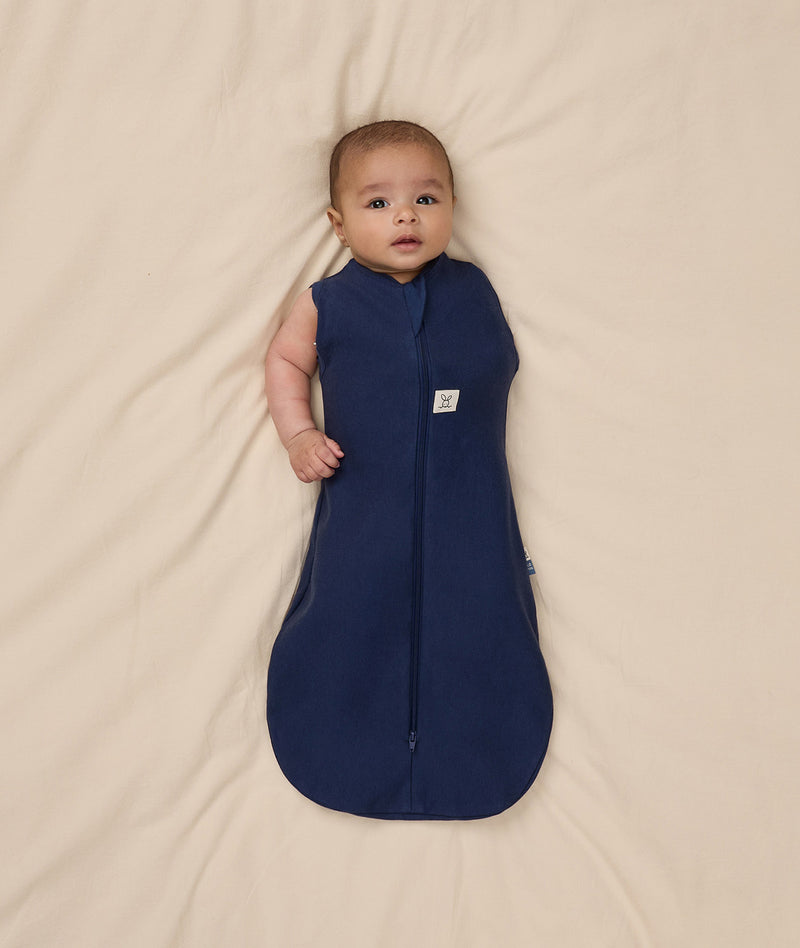 Baby is swaddled in a dark blue ergoPouch Cocoon Swaddle Bag 1.0 TOG with one arm outside.