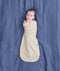 Baby lying on a blue fabric background wearing a cream patterned Cocoon Swaddle Bag with one hand out.