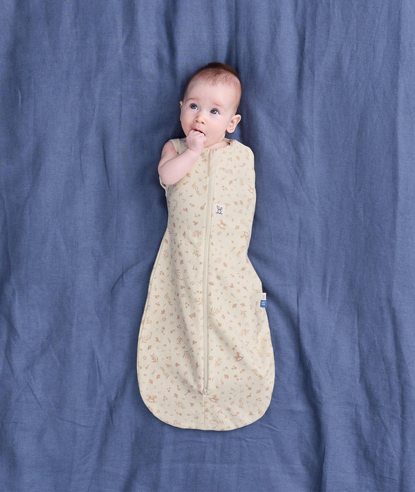 Baby lying on a blue fabric background wearing a cream patterned Cocoon Swaddle Bag with one hand out.