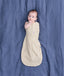 Baby lying on a blue fabric background wearing a cream patterned Cocoon Swaddle Bag with one hand out.