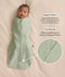 Baby swaddled in a green floral Cocoon Swaddle Bag, lying on a cream-coloured blanket with text callouts highlighting organic cotton and two-way zip features.