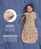 Baby lying on a blue fabric background wearing a tan patterned Cocoon Swaddle Bag with text highlighting press studs and stretchy & bell-shaped features.