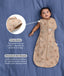 Baby lying on a blue fabric background wearing a tan patterned Cocoon Swaddle Bag with text highlighting press studs and stretchy & bell-shaped features.