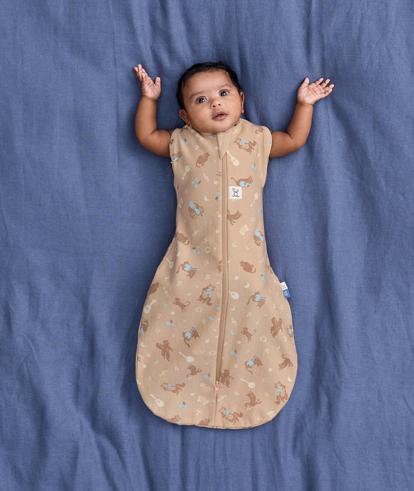 Baby lying on a blue fabric background wearing a tan patterned Cocoon Swaddle Bag with both arms out.