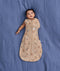 Baby lying on a blue fabric background wearing a tan patterned Cocoon Swaddle Bag with one hand out.