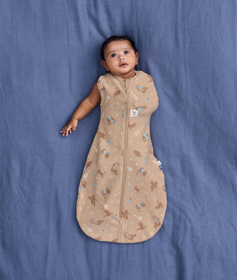 Baby lying on a blue fabric background wearing a tan patterned Cocoon Swaddle Bag with one hand out.