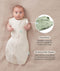 Baby lying on a beige fabric in an oatmeal-coloured swaddle bag with arms out, featuring callout text highlighting press studs and a bell-shaped design.