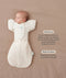Baby lying on a beige surface in an oatmeal-colored swaddle bag paired with a Butterfly Cardi sleeve attachment, featuring product text callouts highlighting hands up sleeping and wash and wear care.