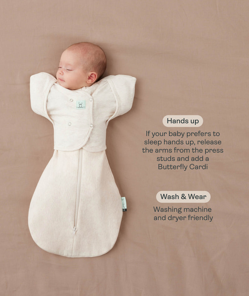 Baby lying on a beige surface in an oatmeal-colored swaddle bag paired with a Butterfly Cardi sleeve attachment, featuring product text callouts highlighting hands up sleeping and wash and wear care.
