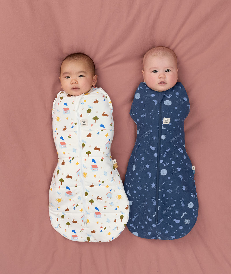 Two babies lying on a pink surface wearing ergoPouch Cocoon Swaddle Bags. One wearing a 2.5 TOG Swaddle Bag in Daydream, while the other wearing a 1.0 TOG Swaddle Bag in Starbeam
