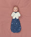 Baby lying on a soft pink surface, wearing a blue Cocoon Swaddle Bag with a moon-and-star print and a cream Butterfly Cardi, looking up toward the camera.