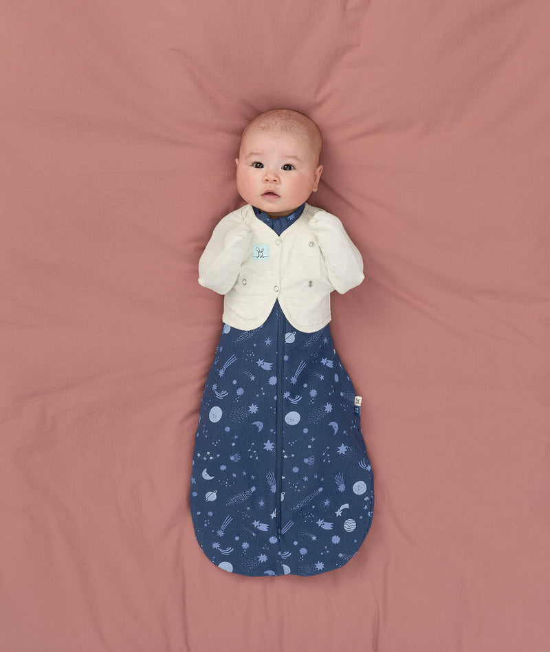 Baby lying on a soft pink surface, wearing a blue Cocoon Swaddle Bag with a moon-and-star print and a cream Butterfly Cardi, looking up toward the camera.