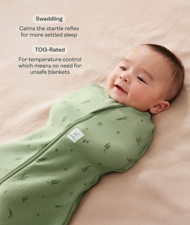 Smiling baby lying on a beige surface being swaddled in an ergoPouch Cocoon Swaddle Bag Willow with text highlighting swaddling benefit and TOG rating feature.