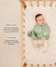 Baby lying in a wooden cot wearing a sage green patterned swaddle bag over footed layer, with product feature text callouts highlighting organic cotton fabric and two-way zip features.