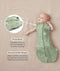 Baby lying on a beige surface in a green patterned swaddle bag with arms out, featuring callout text highlighting press studs and a bell-shaped design.