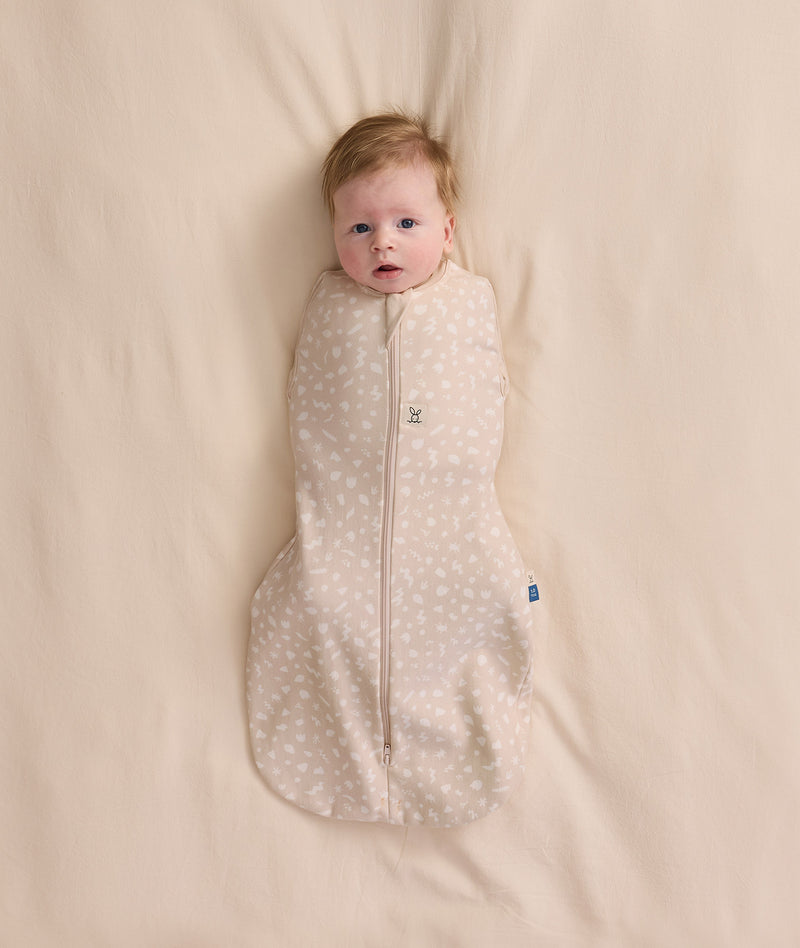 An overhead view of a baby swaddled in a patterned beige Cocoon Swaddle Bag, lying flat on a cream-colored blanket.