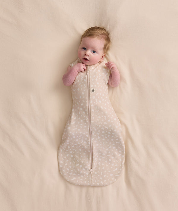 Baby lying on a cream blanket, wearing a patterned Cocoon Swaddle Bag with both arms out