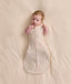 Baby lying on a cream blanket, wearing a patterned Cocoon Swaddle Bag with both arms out