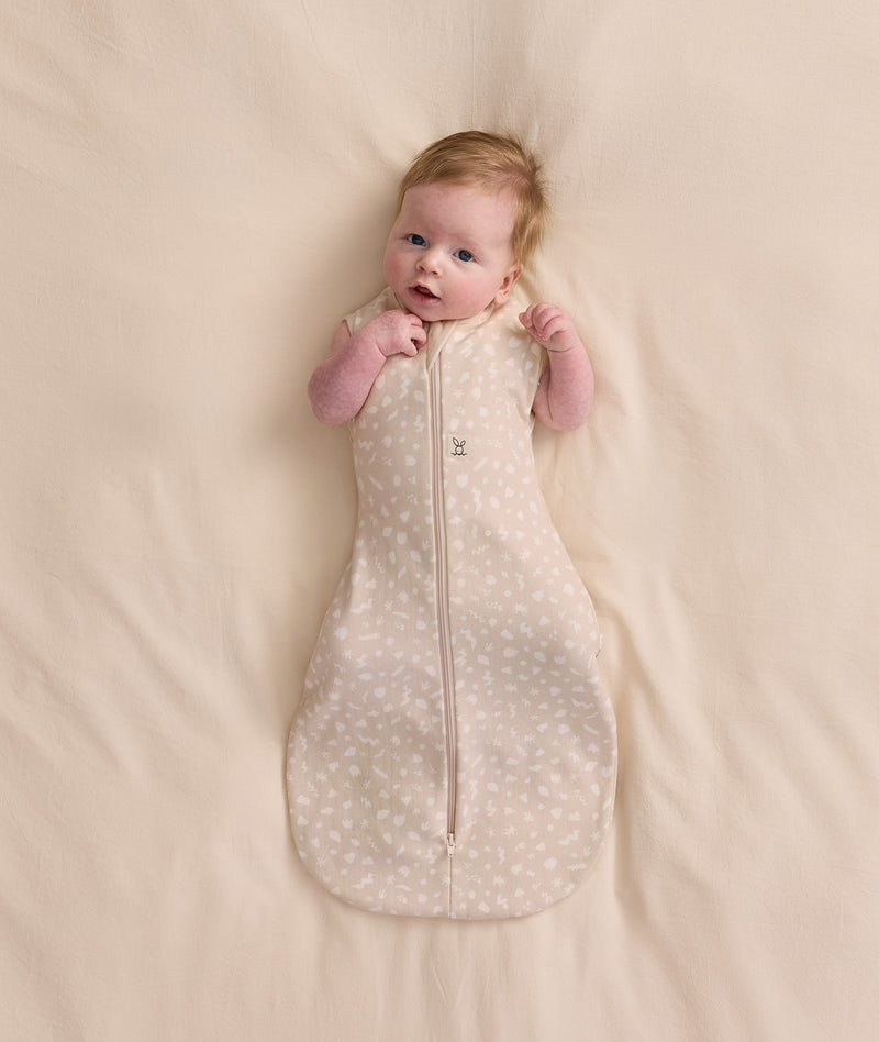 Baby lying on a cream blanket, wearing a patterned Cocoon Swaddle Bag with both arms out