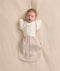 An overhead view of a baby lying flat on a cream-colored sheet, wearing a patterned beige Cocoon Swaddle Bag with a Butterfly Cardi layered over top.