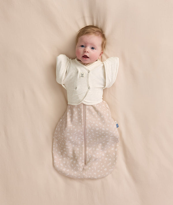 An overhead view of a baby lying flat on a cream-colored sheet, wearing a patterned beige Cocoon Swaddle Bag with a Butterfly Cardi layered over top.