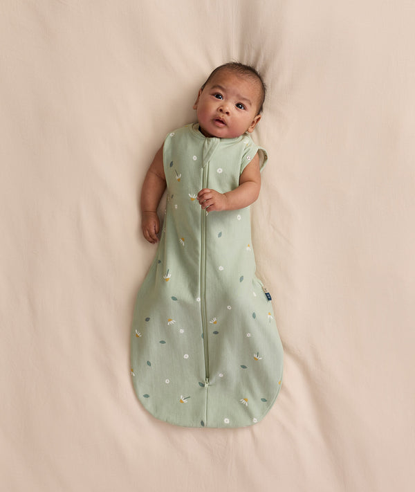 Baby lying on a cream blanket, wearing an ergoPouch Cocoon Swaddle Bag 1.0 TOG with both arms out.