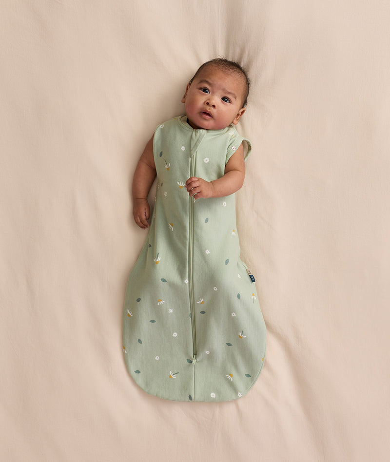 Baby lying on a cream blanket, wearing an ergoPouch Cocoon Swaddle Bag 1.0 TOG with both arms out.
