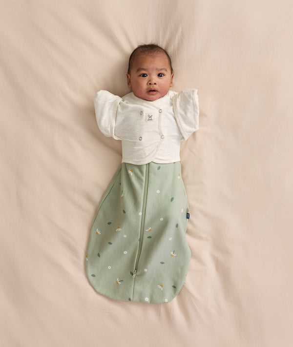 Baby lying on a cream -coloured blanket, wearing a sage green floral-patterned Cocoon Swaddle Bag layered with a Butterfly Cardi over top