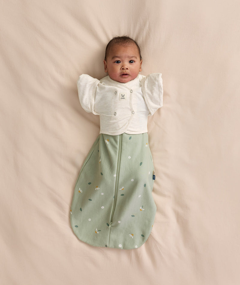Baby lying on a cream -coloured blanket, wearing a sage green floral-patterned Cocoon Swaddle Bag layered with a Butterfly Cardi over top