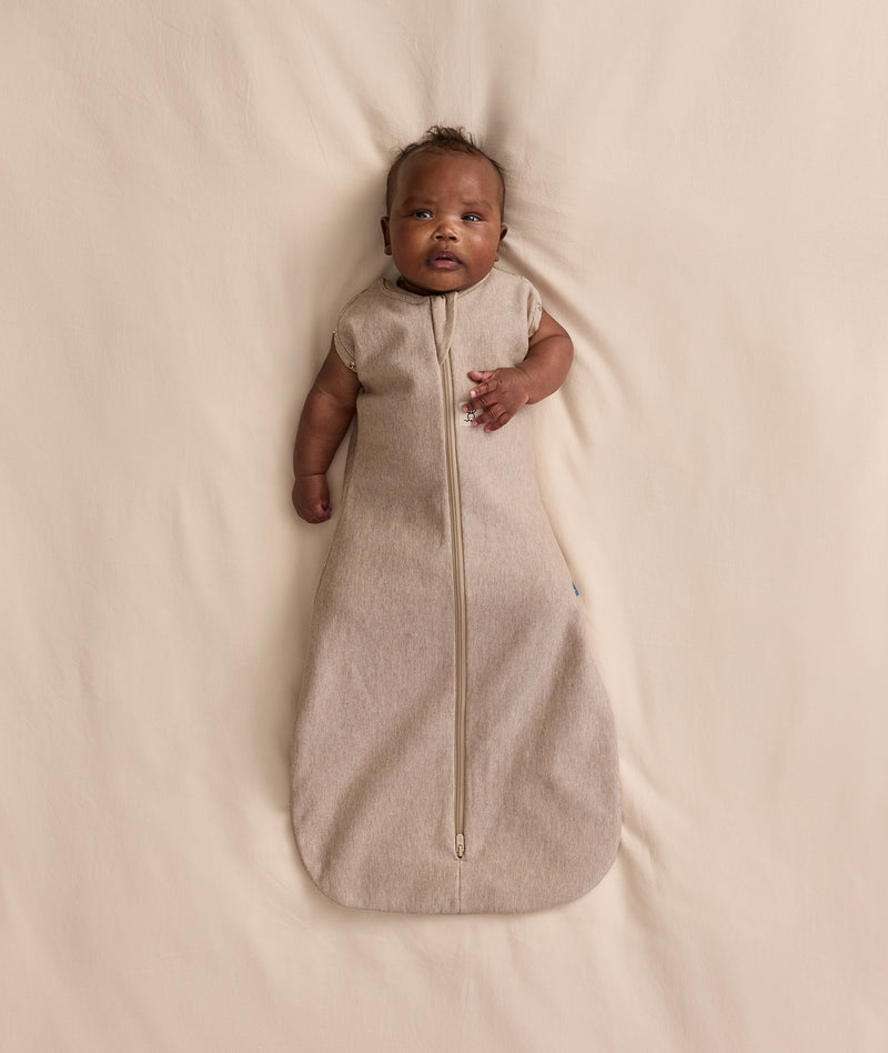 Baby lying on a cream blanket, wearing an ergoPouch Cocoon Swaddle Bag with both arms out