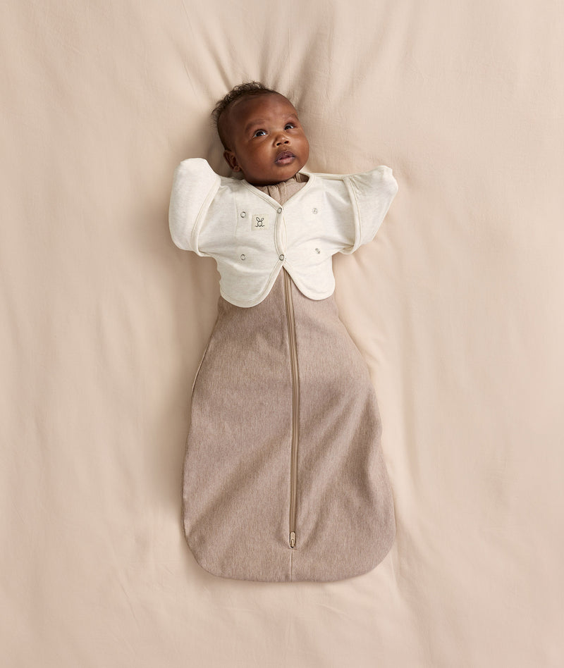 Baby lying on a cream-coloured blanket wearing a Cocoon Swaddle Bag layered with a Butterfly Cardi over top.