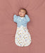 Baby lying on a pink surface, dressed in a white Cocoon Swaddle Bag with a colourful print and wearing a blue Butterfly Cardi over it.
